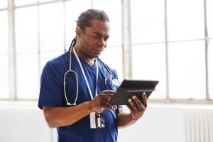 Black male doctor with stethoscope and in blue scrubs looks at tablet