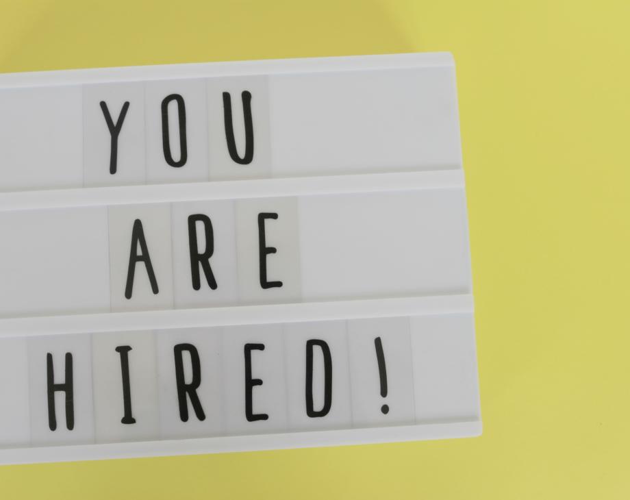 Against a yellow background reader board of black text that says, "YOU ARE HIRED!"
